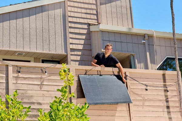 A man is installing a solar panel on a fence while standing on a deck, surrounded by greenery and a residential building.