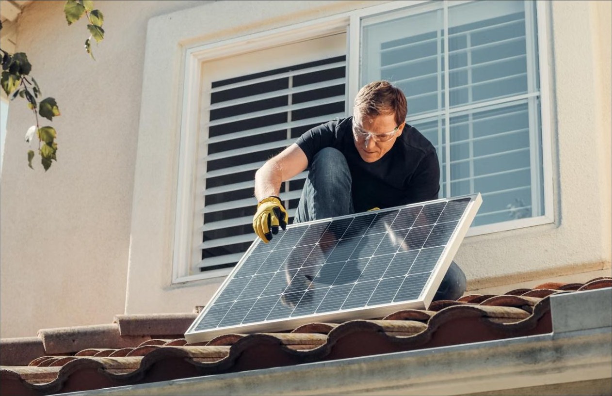 A man in work gloves and safety glasses setting up a compact solar panel in bright sun outside his window
